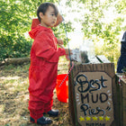 A young child wearing a red waterproof suit, standing at a mud pie stand on a sunny day, looking off to the side.